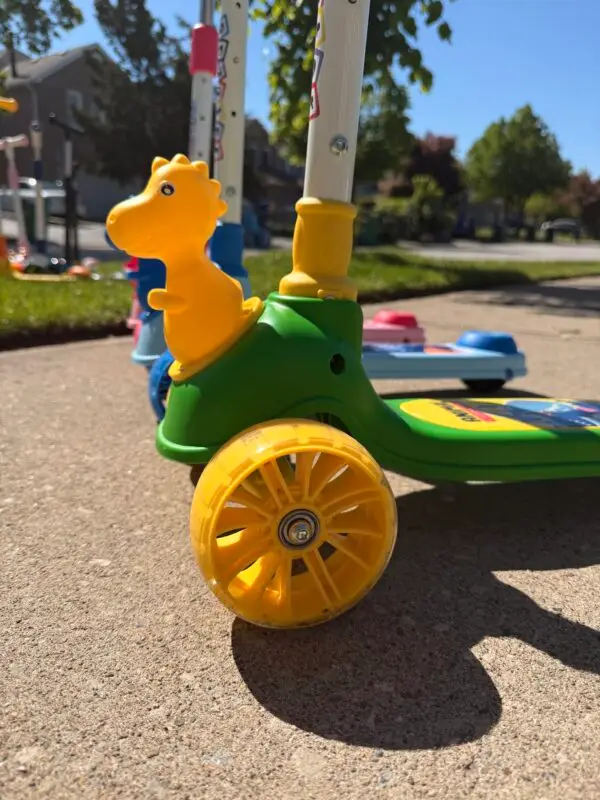 Close-up of a yellow dinosaur head attachment on a green Giant Galaxi 3-wheel scooter with blue and red models blurred in the background.