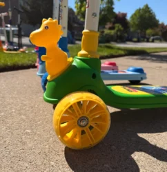 Close-up of a yellow dinosaur head attachment on a green Giant Galaxi 3-wheel scooter with blue and red models blurred in the background.