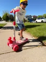 Young boy riding a pink Giant Galaxi Dino series 3-wheel scooter with a dinosaur head attachment on a sunny sidewalk.