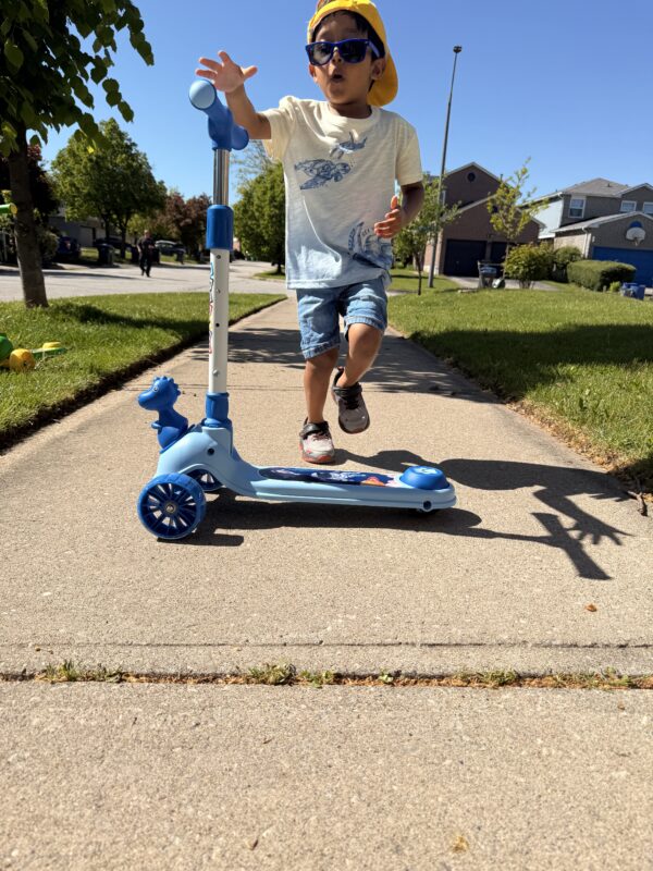 A young boy in a yellow hat and sunglasses riding a blue Giant Galaxi Dino series kick scooter on a sunny sidewalk.