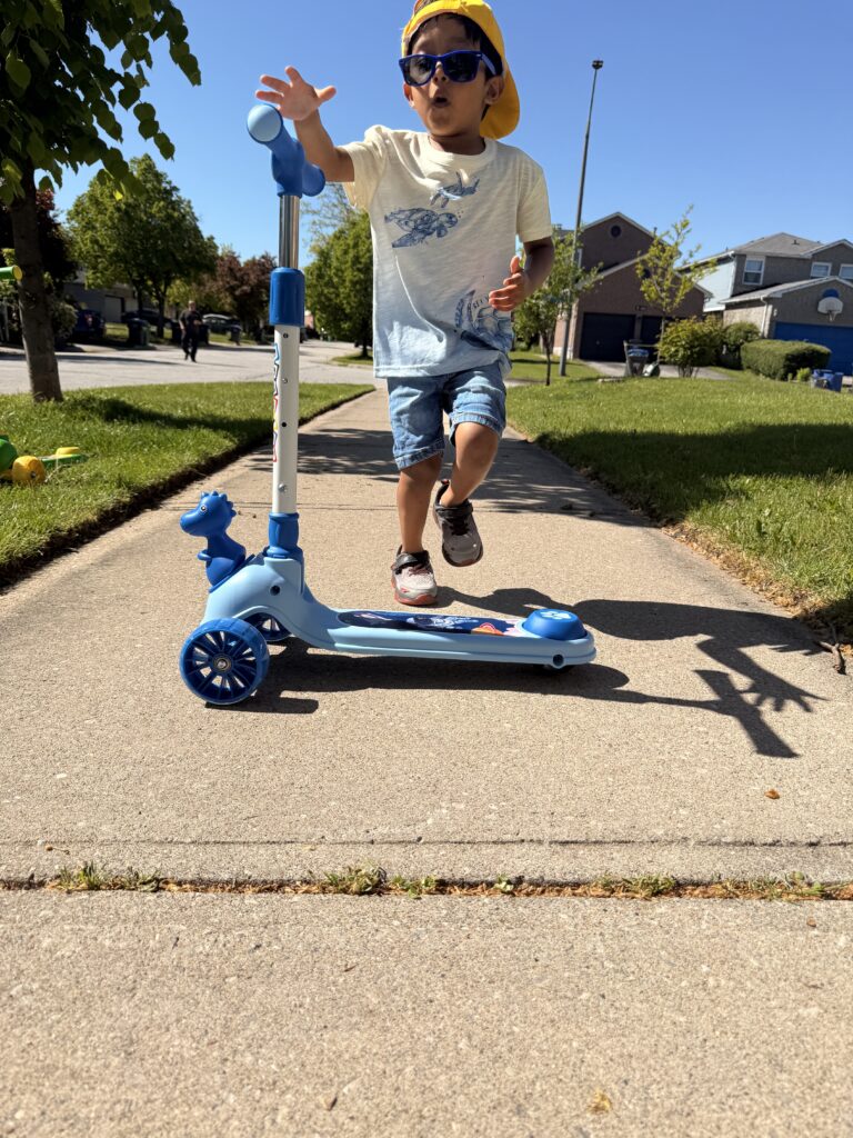 A young boy in a yellow hat and sunglasses riding a blue Giant Galaxi Dino series kick scooter on a sunny sidewalk.