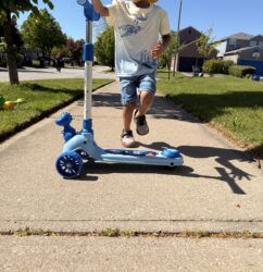 A young boy in a yellow hat and sunglasses riding a blue Giant Galaxi Dino series kick scooter on a sunny sidewalk.